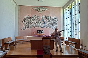 Mann spielt Gitarre in einer hellen Kapelle mit Holzbankreihen und einem großen Wandbild mit mehreren Figuren hinter dem Altar