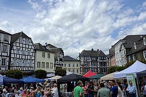 Marktplatz mit Fachwerkhäusern und mehreren Menschen, die an Ständen und Tischen unter Zelten und Sonnenschirmen stehen und sitzen