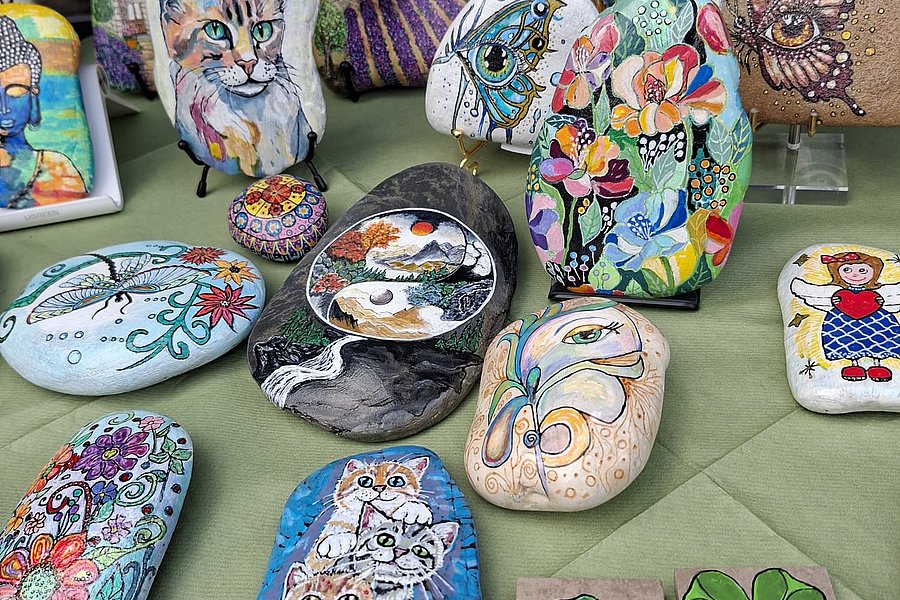 Mehrere bemalte Steine mit Motiven wie Katzen, Schmetterlingen, Blumen, einem Auge und einem Kleeblatt auf einer grünen Oberfläche