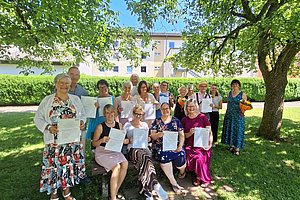 Gruppe von Menschen im Freien unter Bäumen, einige sitzen auf einer Bank, andere stehen, alle halten Papiere in der Hand