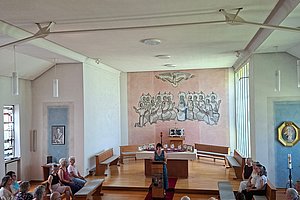 Innenraum einer Kirche mit hölzernen Bänken, einem roten Mittelgang und einer Gruppe von Menschen, die auf den Altarbereich mit einer Wandmalerei blicken